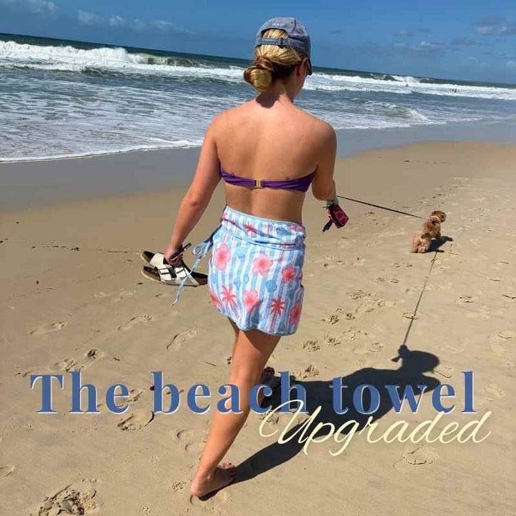 Woman walking on a beach with a dog, wearing a colorful towel.