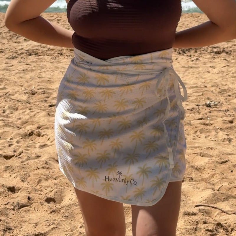 A person standing on a sandy beach wearing a towel wrap with a yellow palm tree pattern and a brown ribbed top. The brand name 'Heavenly Co' is printed near the bottom of the sarong.