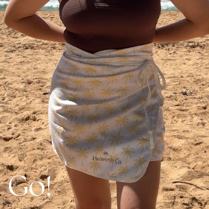 Person wearing a palm tree patterned sarong on a sandy beach with 'Heavenly Co' branding.