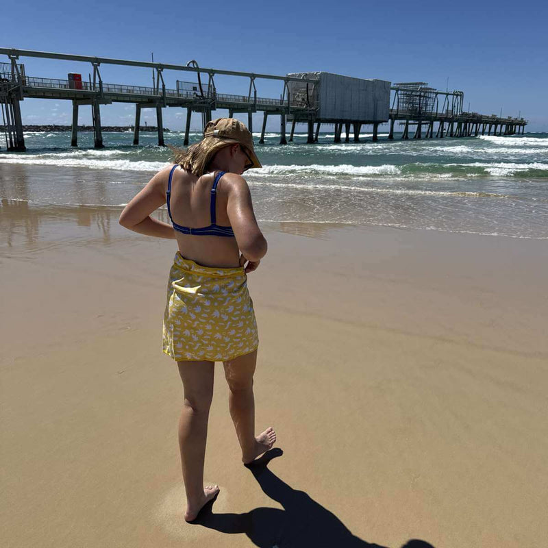 Person in a yellow skirt and blue top standing on a beach with a pier in the background