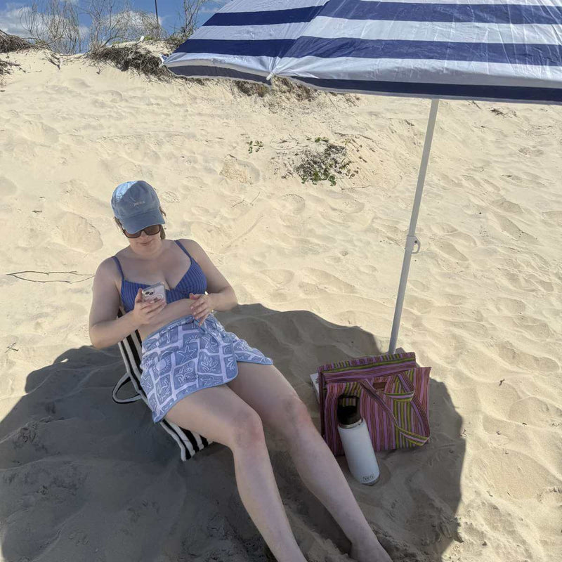 Woman in a blue swimsuit sitting under a striped beach umbrella on sand.