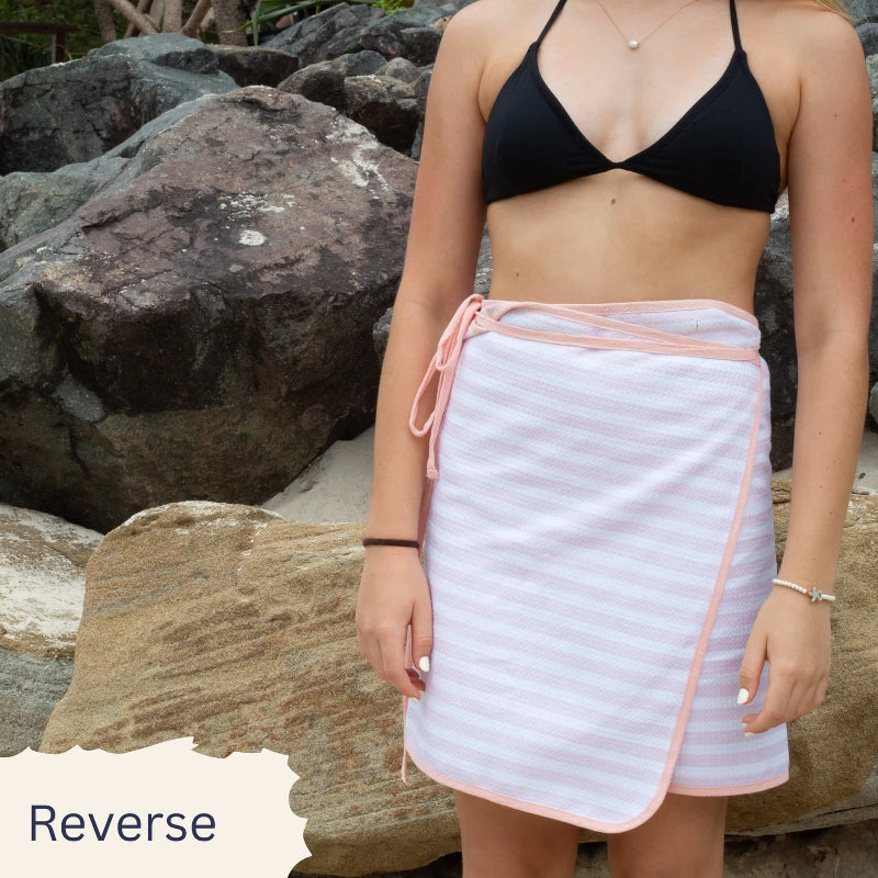 Person wearing a black bikini top and white striped towel with pink trim, standing against rocks.