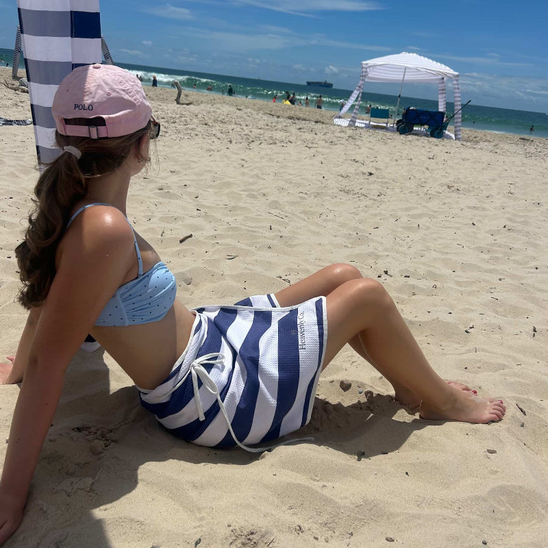 Woman sitting on the beach wearing a blue and white striped skirt and pink cap.