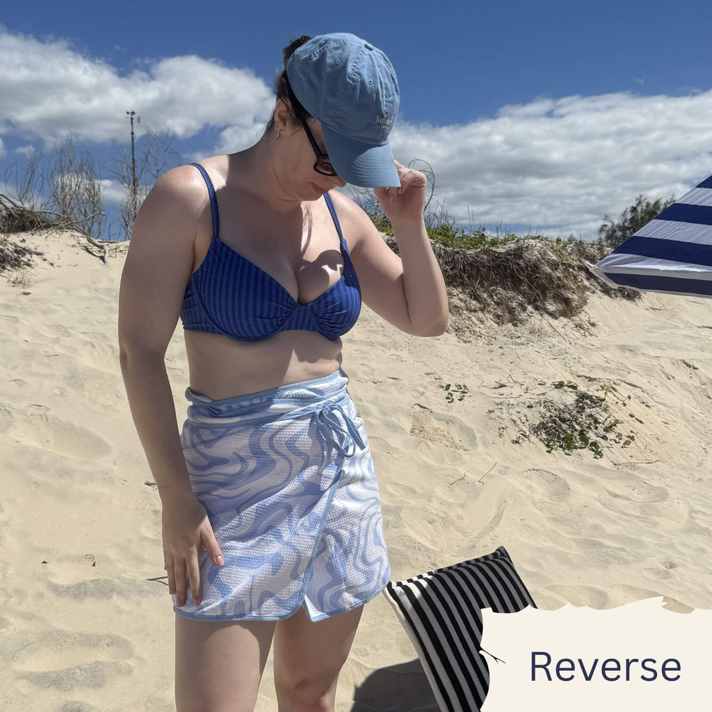 Woman in blue bikini top and patterned shorts standing on a sandy beach with a blue cap.